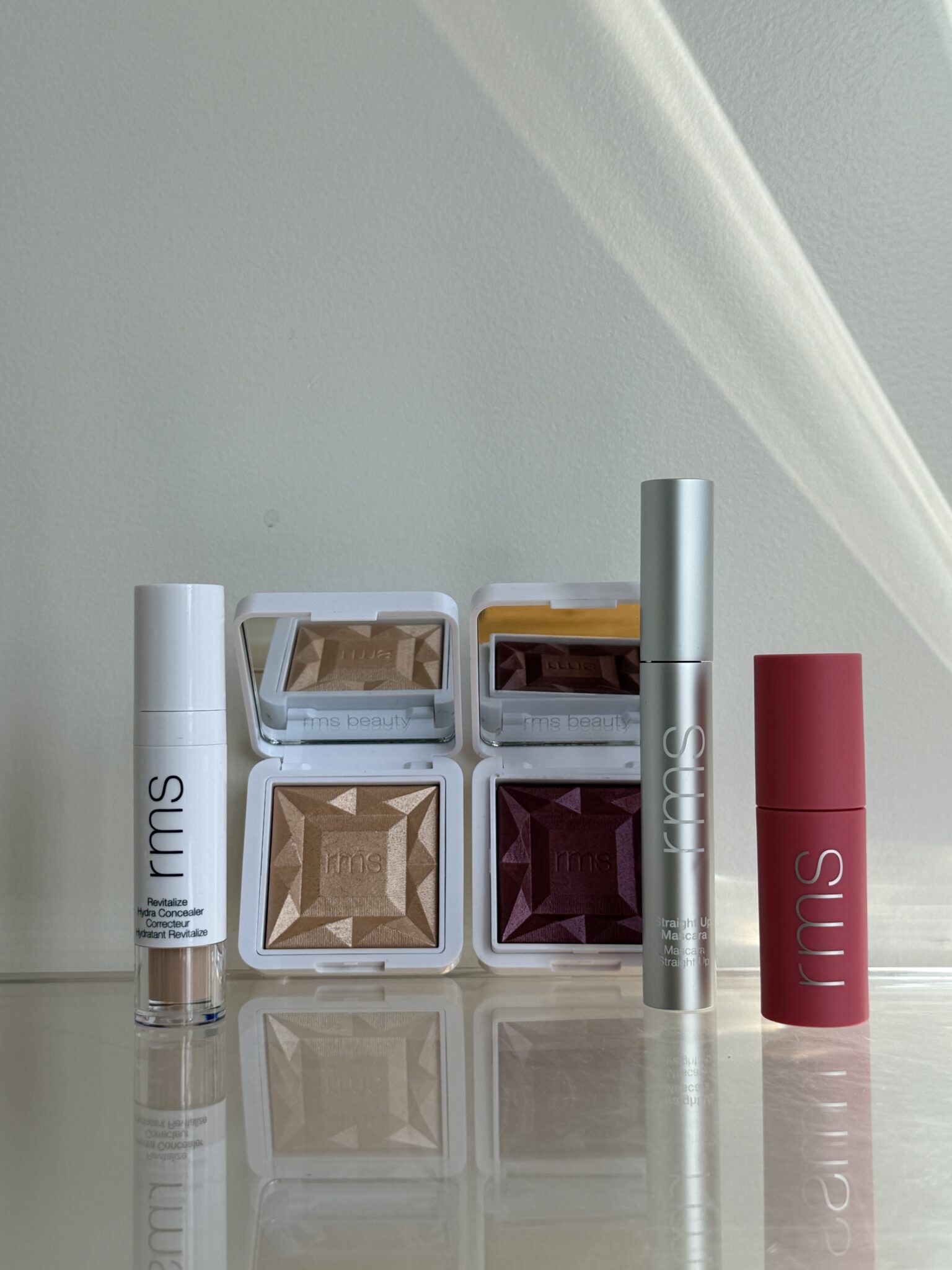 Five RMS Beauty makeup products, including two closed compacts, a mascara, a lipstick, and a liquid concealer, are arranged in a row on a reflective surface against a neutral background.