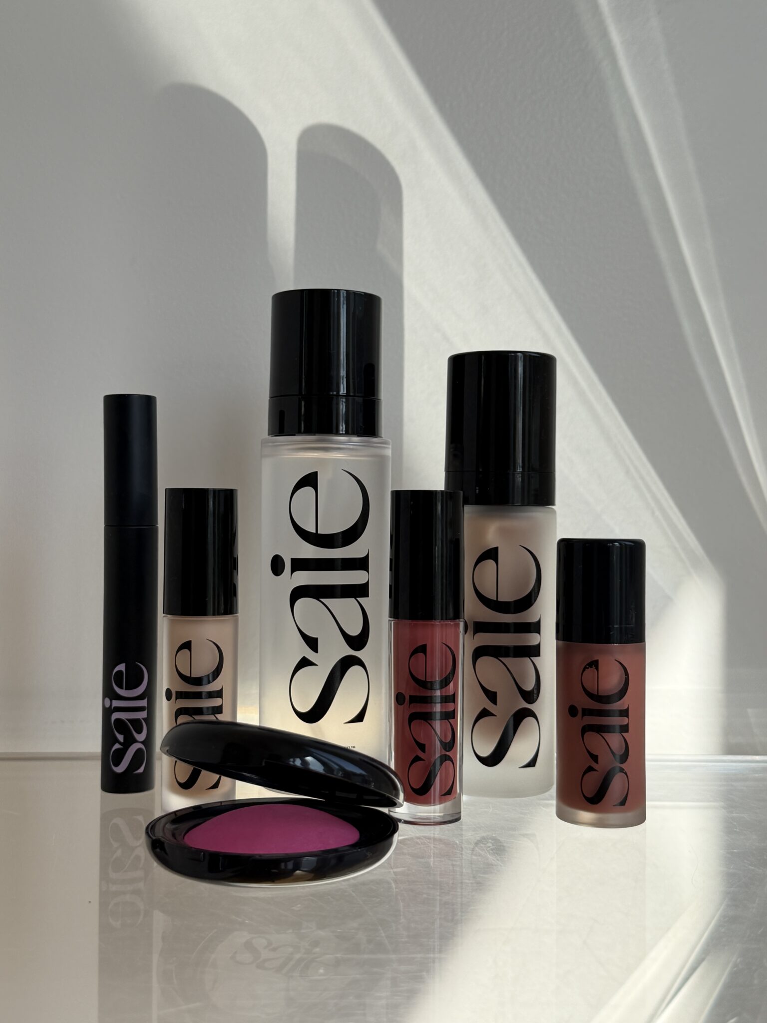 Six Saie makeup products, including foundation, lip gloss, and a compact blush, are arranged on a white surface with light and shadow patterns in the background.