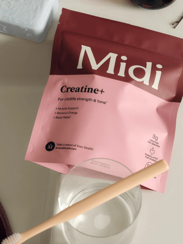 A packet of Midi Creatine+ powder is on a table next to a clear glass of water and a beige toothbrush.