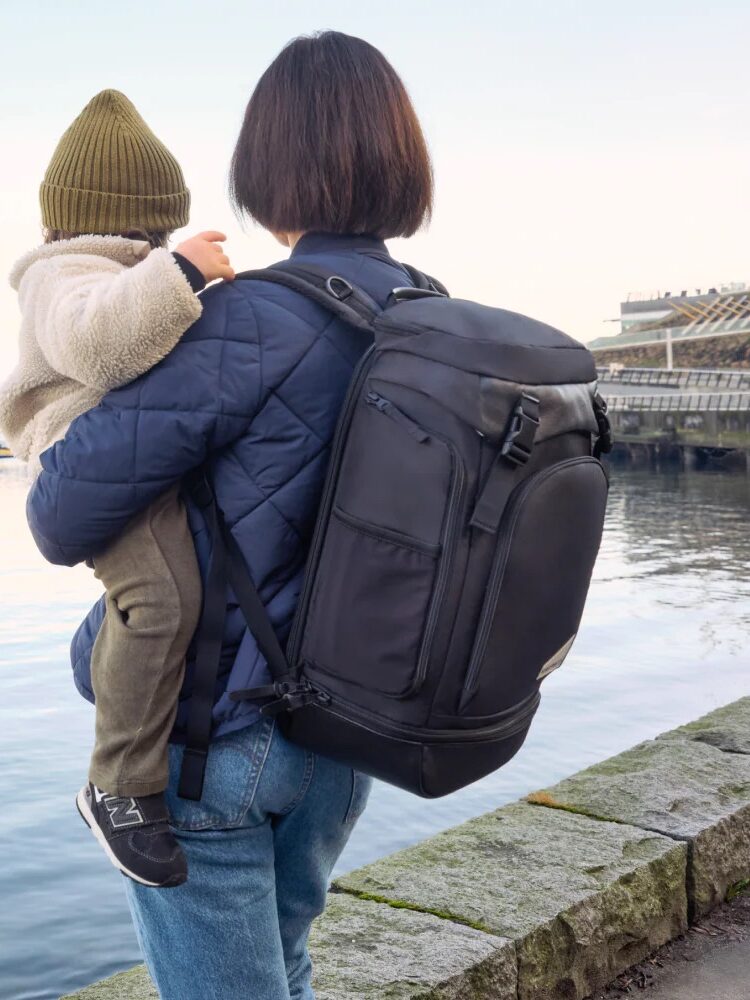 An adult wearing a black backpack holds a child in a beanie and fleece jacket while standing by a waterfront with boats and buildings in the background.