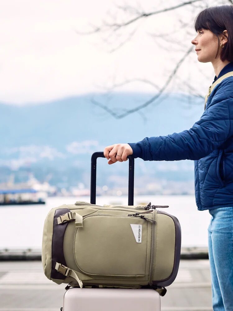 Woman in a blue jacket stands outdoors with two pieces of luggage, one on the ground and one in her hand, with a waterfront and mountains in the background.