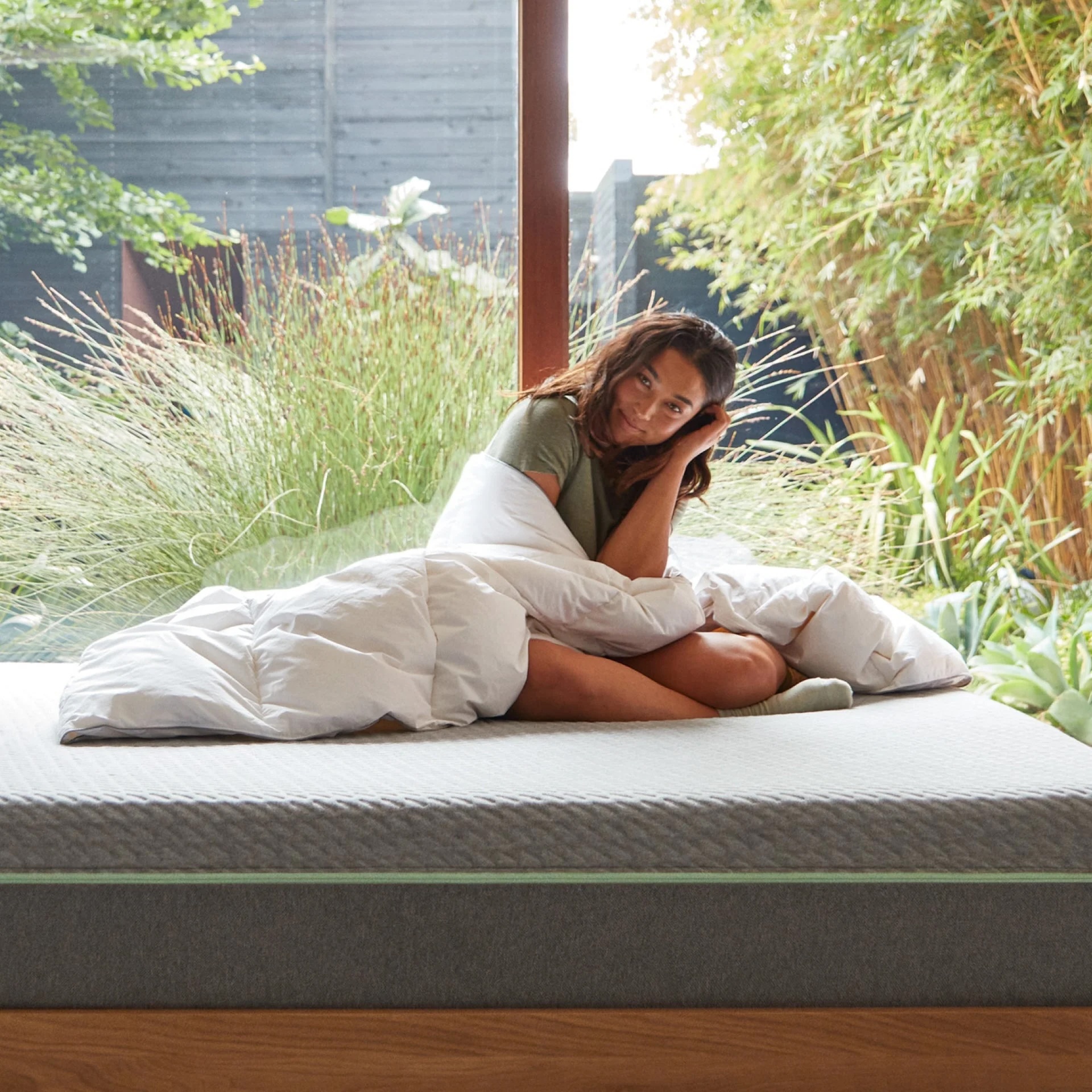 A woman sits on a bed wrapped in a white comforter, next to a large window with plants visible outside.