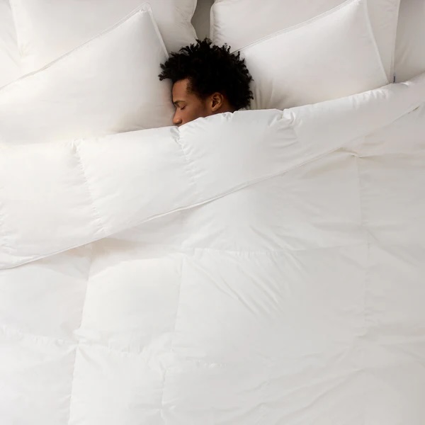 A person is lying in bed mostly covered by a white comforter and white pillows, with only their head visible.