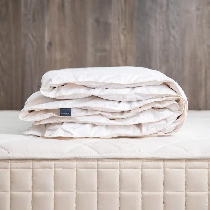 A folded white comforter is placed on top of a beige mattress with a quilted pattern, against a wooden background.