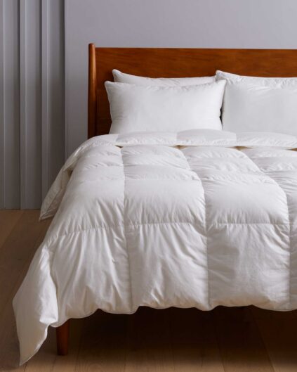 A neatly made bed with white bedding, including a comforter and several pillows, against a wooden headboard in a minimally decorated bedroom.