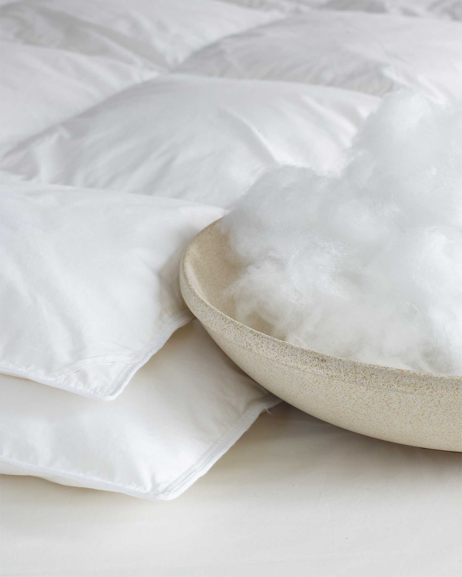 A beige bowl filled with white fluffy filling is placed next to a white quilted pillow or comforter on a light surface.