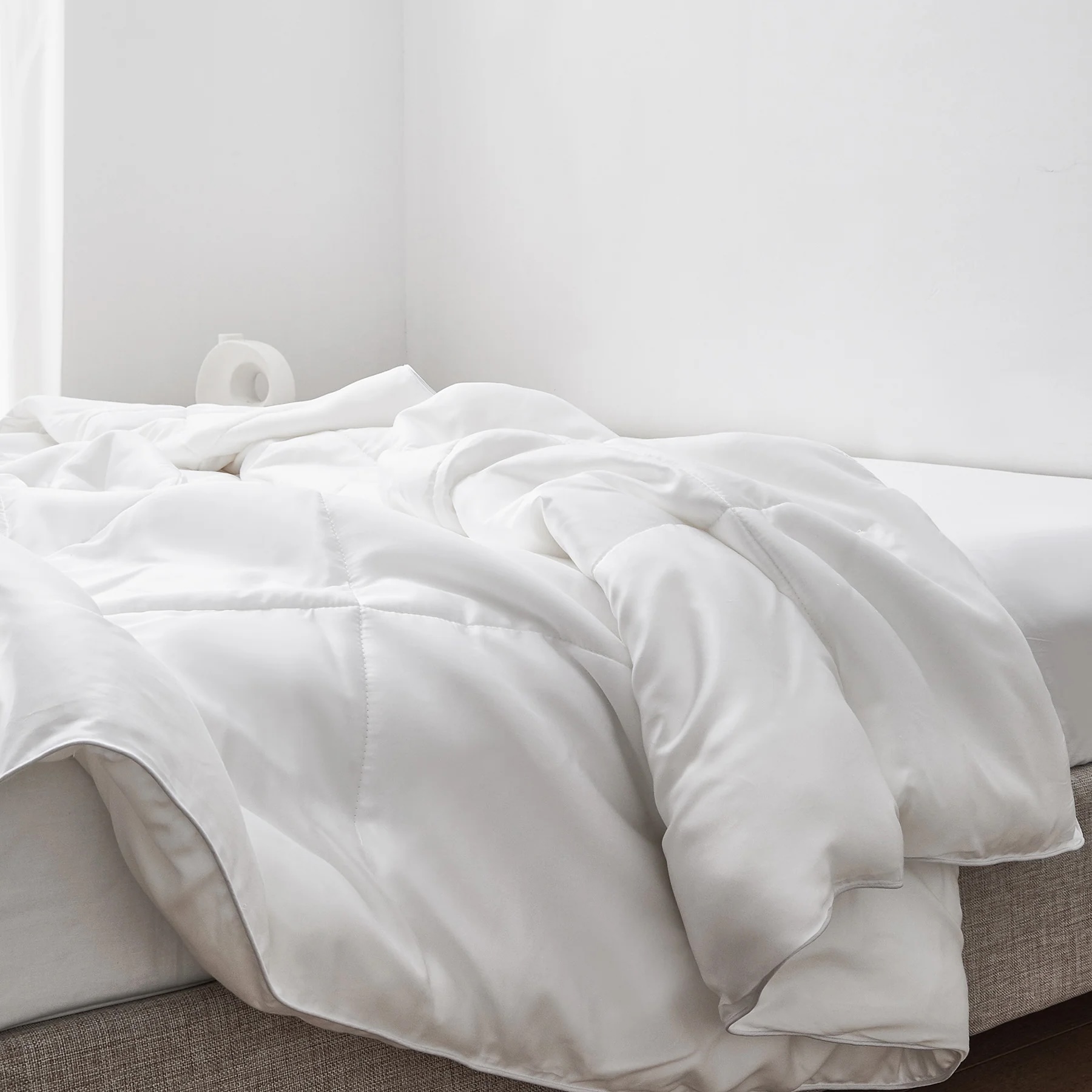 A rumpled white comforter on an unmade bed in a minimal, bright room with white walls and a small white decor item in the background.