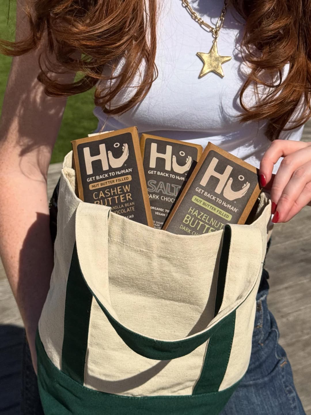 A person with long red hair holds a tote bag containing three Hu chocolate bars: Cashew Butter, Salty Dark Chocolate, and Hazelnut Butter varieties.