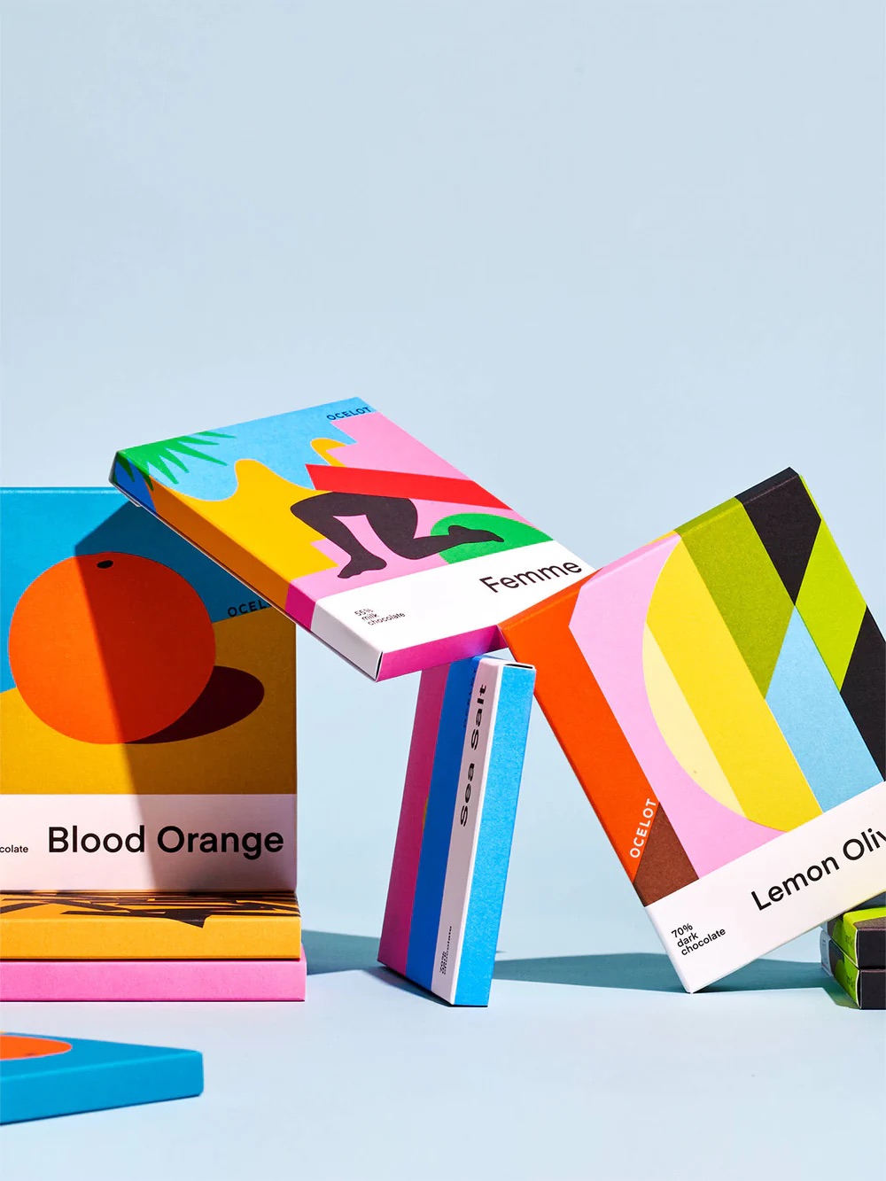 Colorful chocolate bar packages with modern abstract designs are arranged in a balanced, leaning formation against a light blue background.