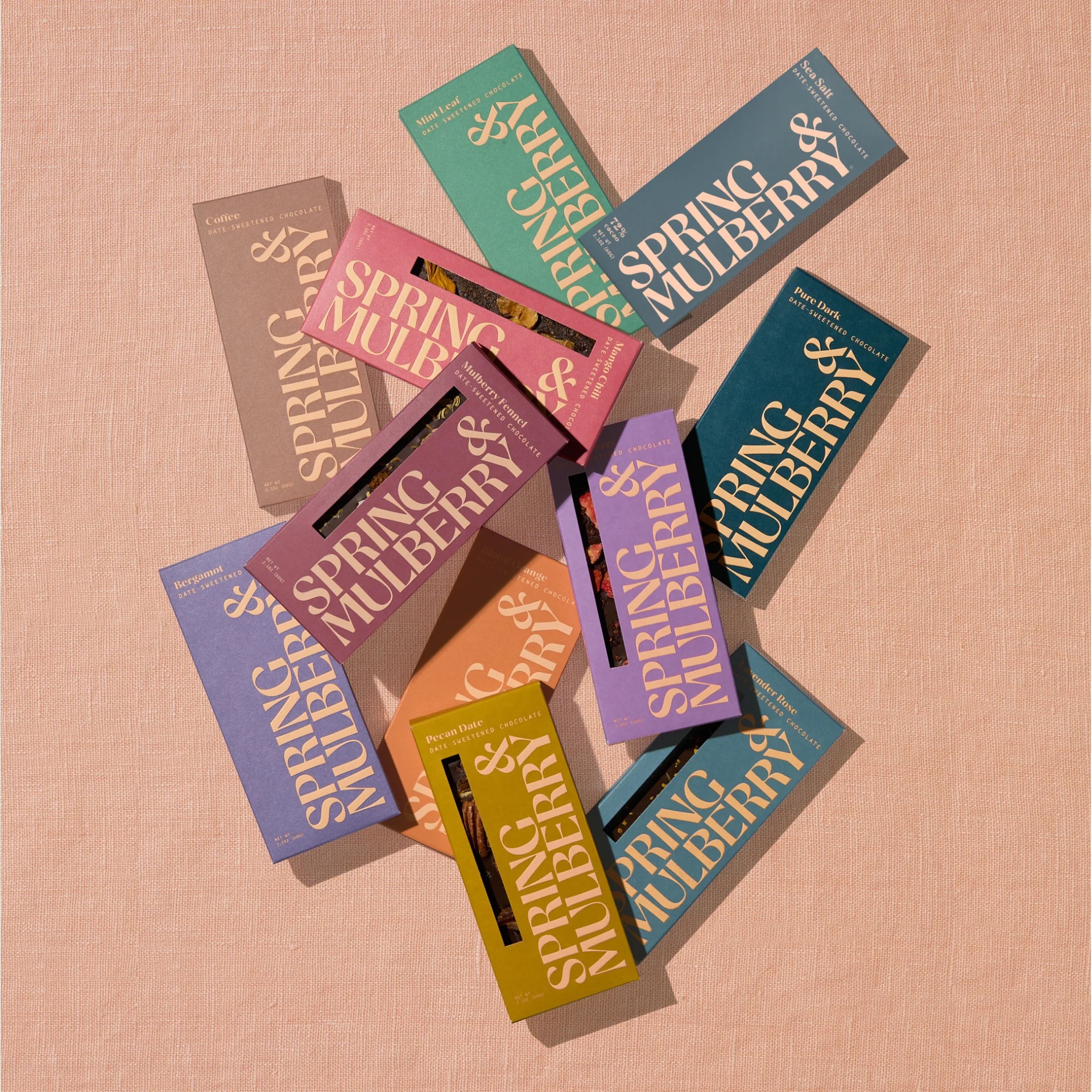 Assorted Spring & Mulberry chocolate bar boxes in various colors are scattered on a peach-colored fabric surface.