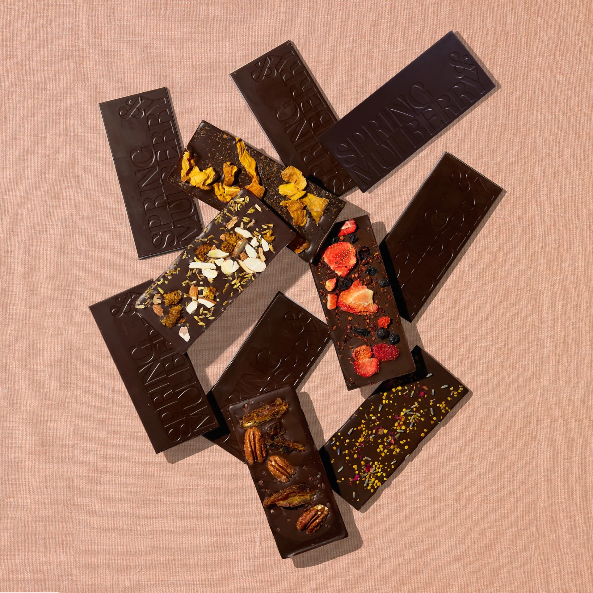 Several dark chocolate bars, some topped with ingredients like nuts, dried fruit, and seeds, are arranged on a light textured surface.