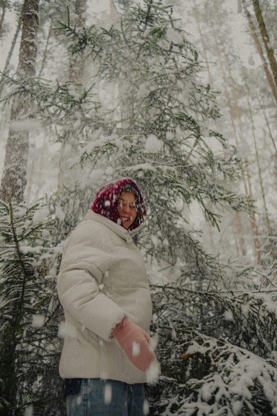 A person in a white winter coat and red hat stands in a snowy forest as large snowflakes fall around them.