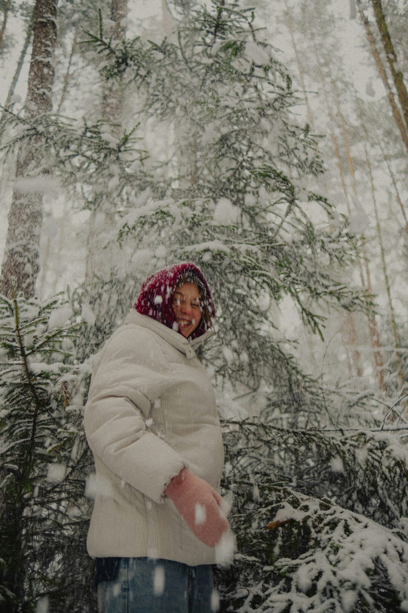 A person in a white winter coat and red hat stands in a snowy forest as large snowflakes fall around them.