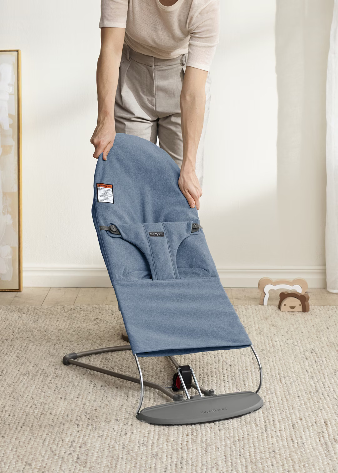 A person is adjusting a blue baby bouncer chair on a carpeted floor in a softly lit room.