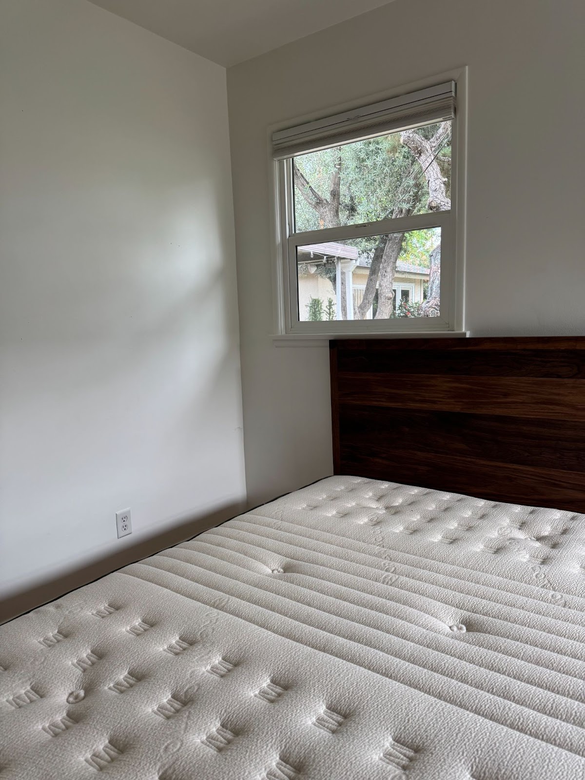 A bare mattress with a wooden headboard is positioned near a window in a minimally furnished room with white walls. Trees and a house are visible outside the window.