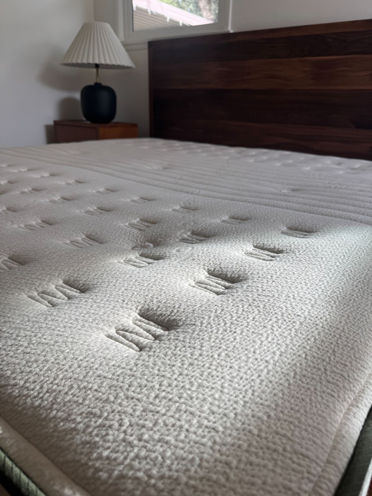 Close-up of a mattress with a textured surface on a bed frame, with a lamp and nightstand in the background.