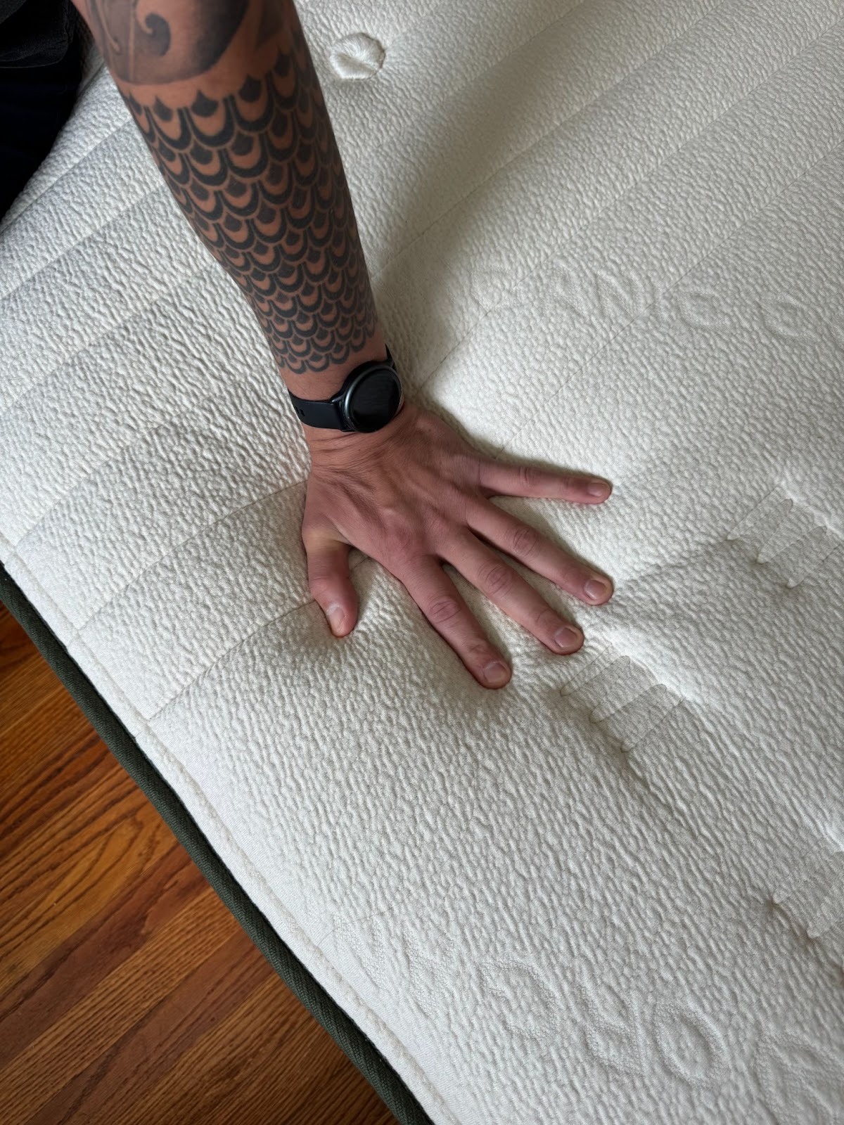 A person's hand with a smartwatch presses down on a textured mattress, showing its softness and indentation.
