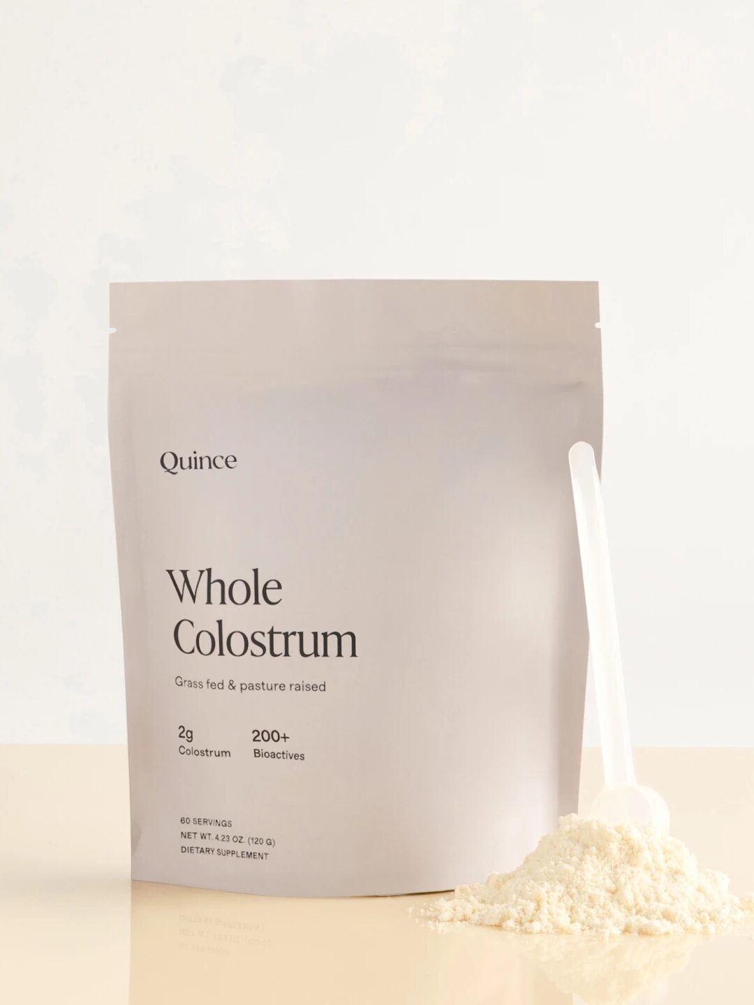 A pouch labeled "Quince Whole Colostrum" stands upright next to a scoop of white powder on a light surface.