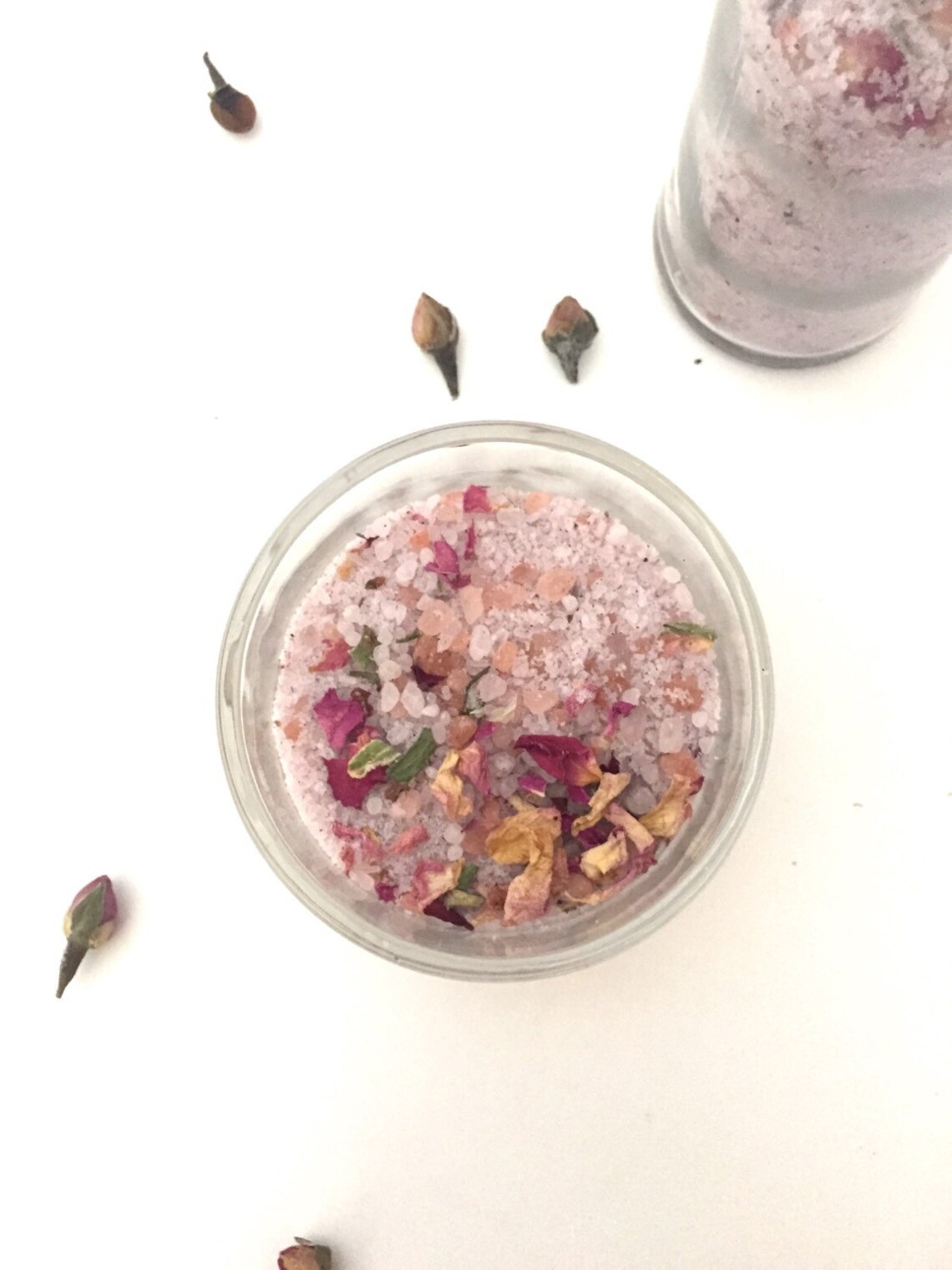 A glass jar filled with pink bath salts and dried flower petals, with a few flower buds scattered on a white surface.