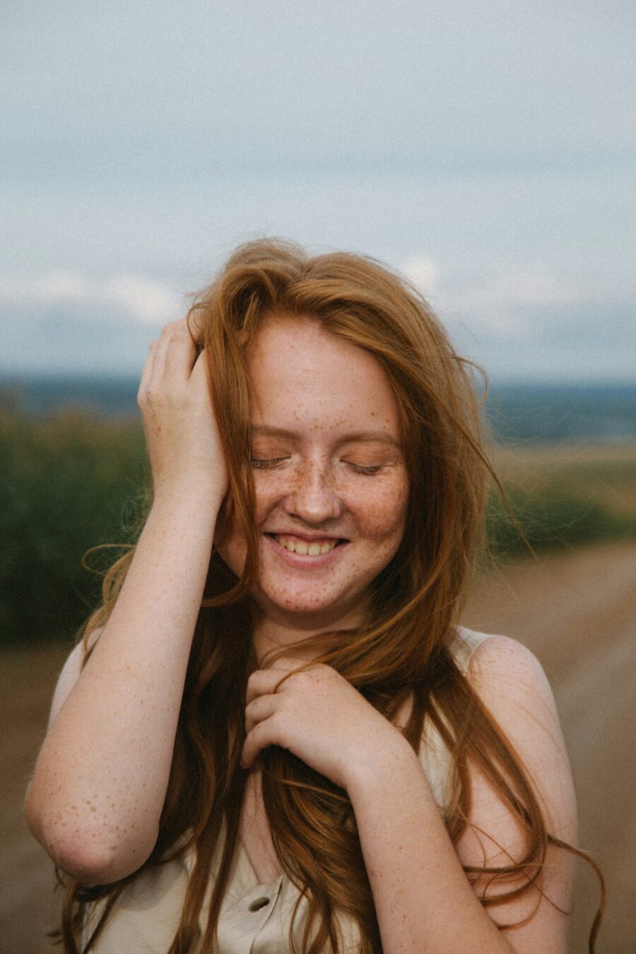 A person with long red hair and freckles stands outdoors on a dirt road, smiling with eyes closed and one hand touching their hair.