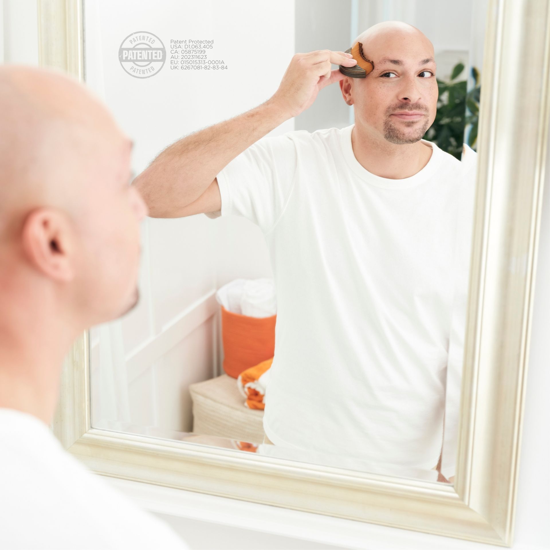 A bald man in a white t-shirt uses a brush on his scalp while looking at himself in a bathroom mirror.