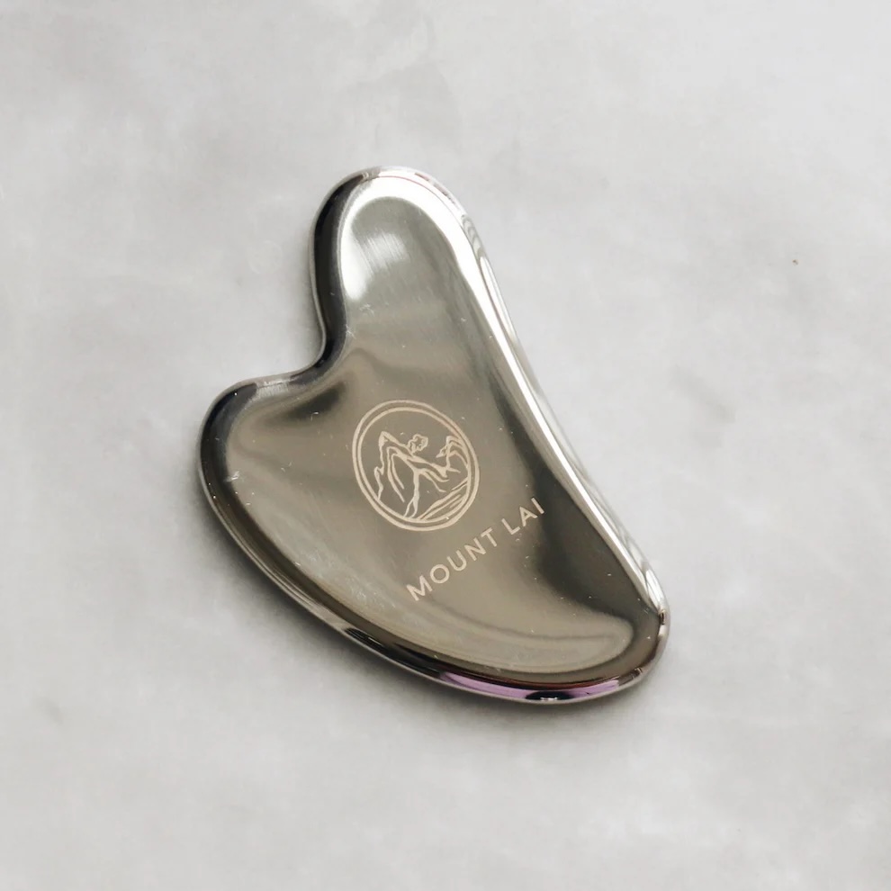 A stainless steel gua sha tool with the Mount Lai logo, placed on a light gray marble surface.