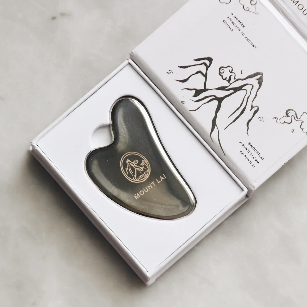 A metal Mount Lai gua sha tool with engraved logo rests in a white box, next to an illustrated instruction card.