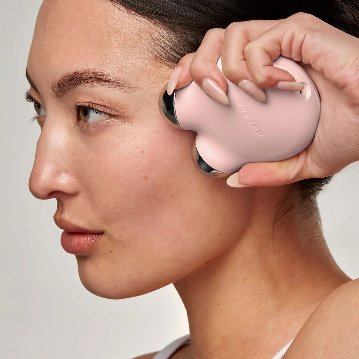 A person uses a pink handheld facial device on their cheek, holding it with one hand close to their face.