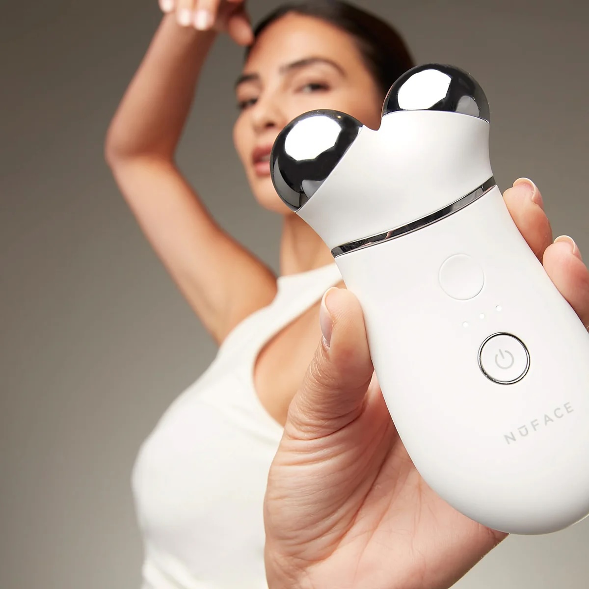 A person holds a white NuFACE facial toning device with two metallic spheres, while another person poses in the background.
