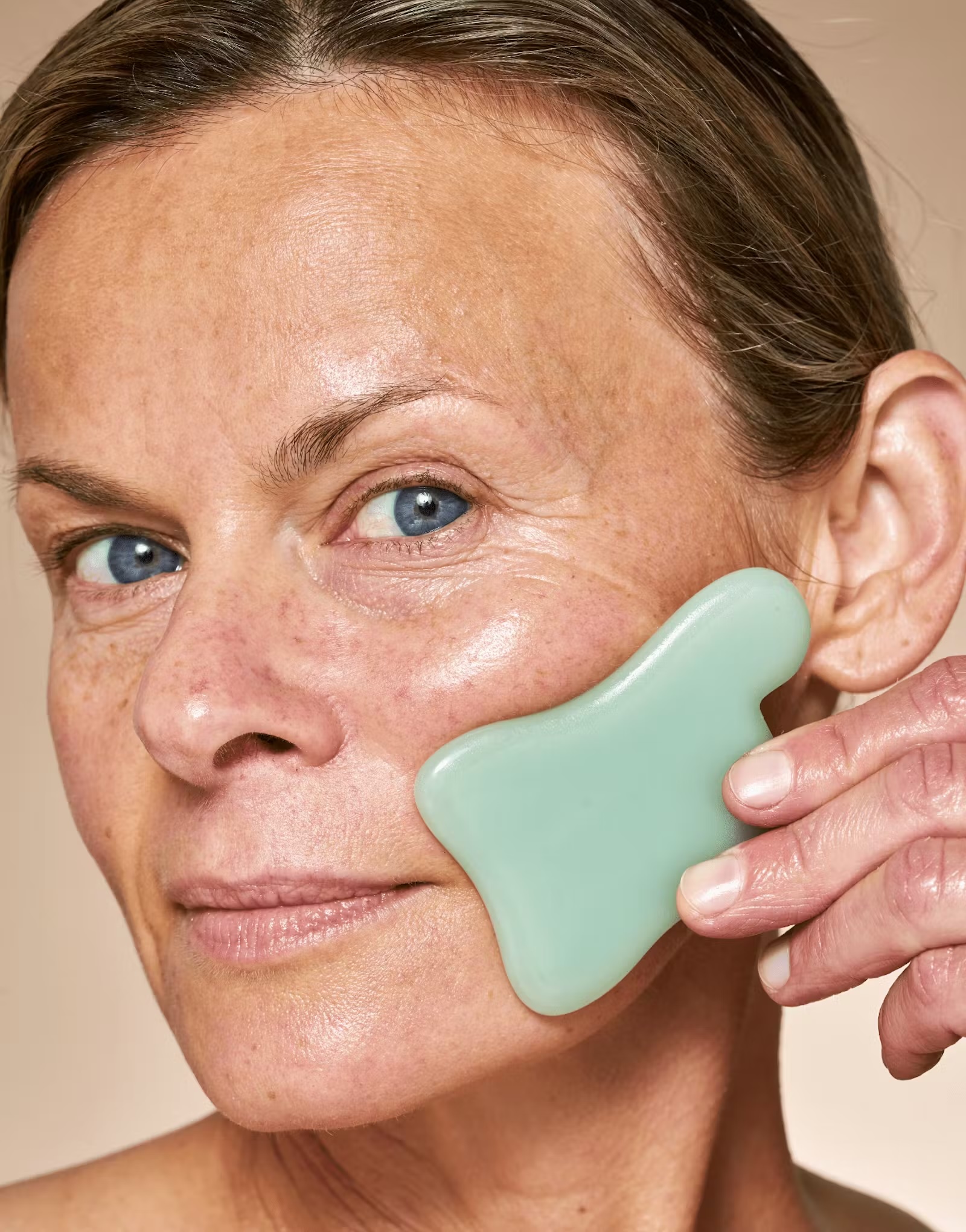 A person uses a green gua sha facial tool to massage their cheek, highlighting smooth, healthy skin and subtle facial lines.