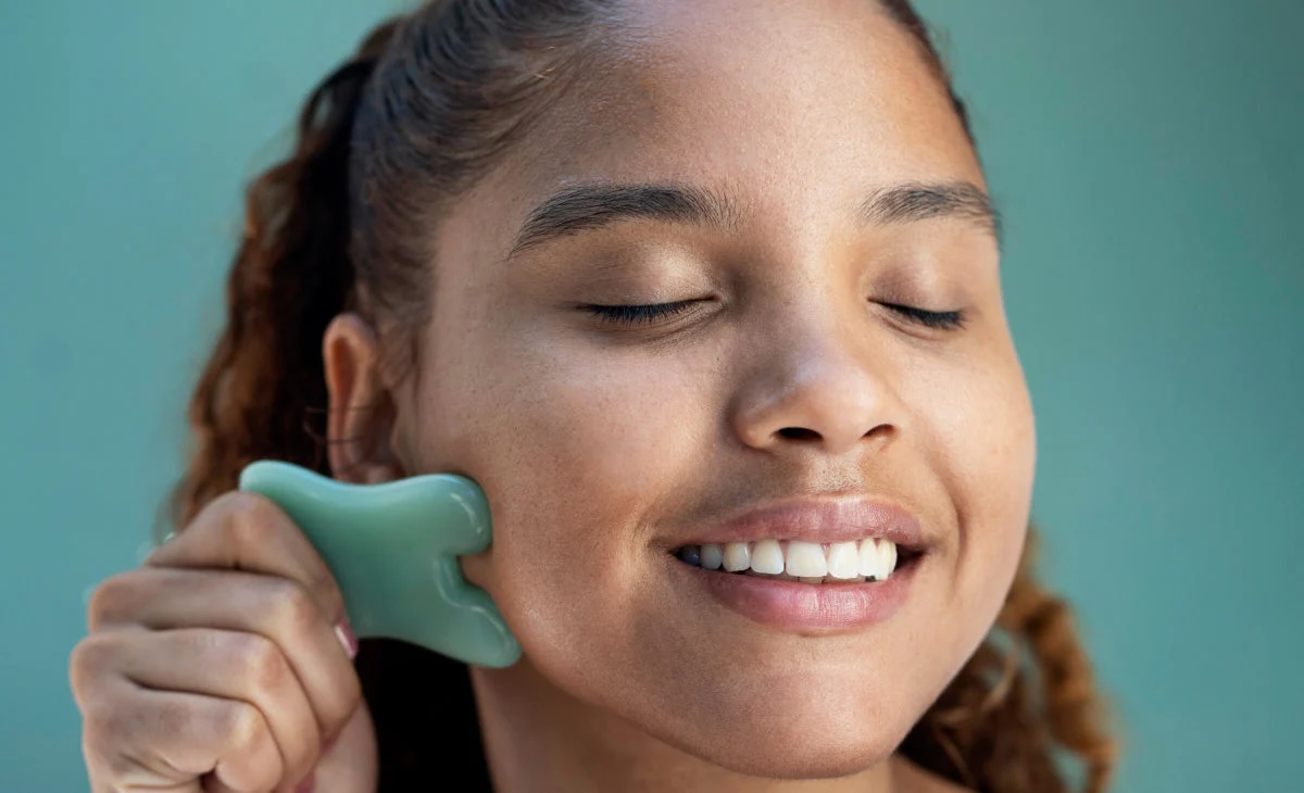 A woman with closed eyes uses a jade gua sha tool to massage her cheek, smiling slightly against a plain teal background.