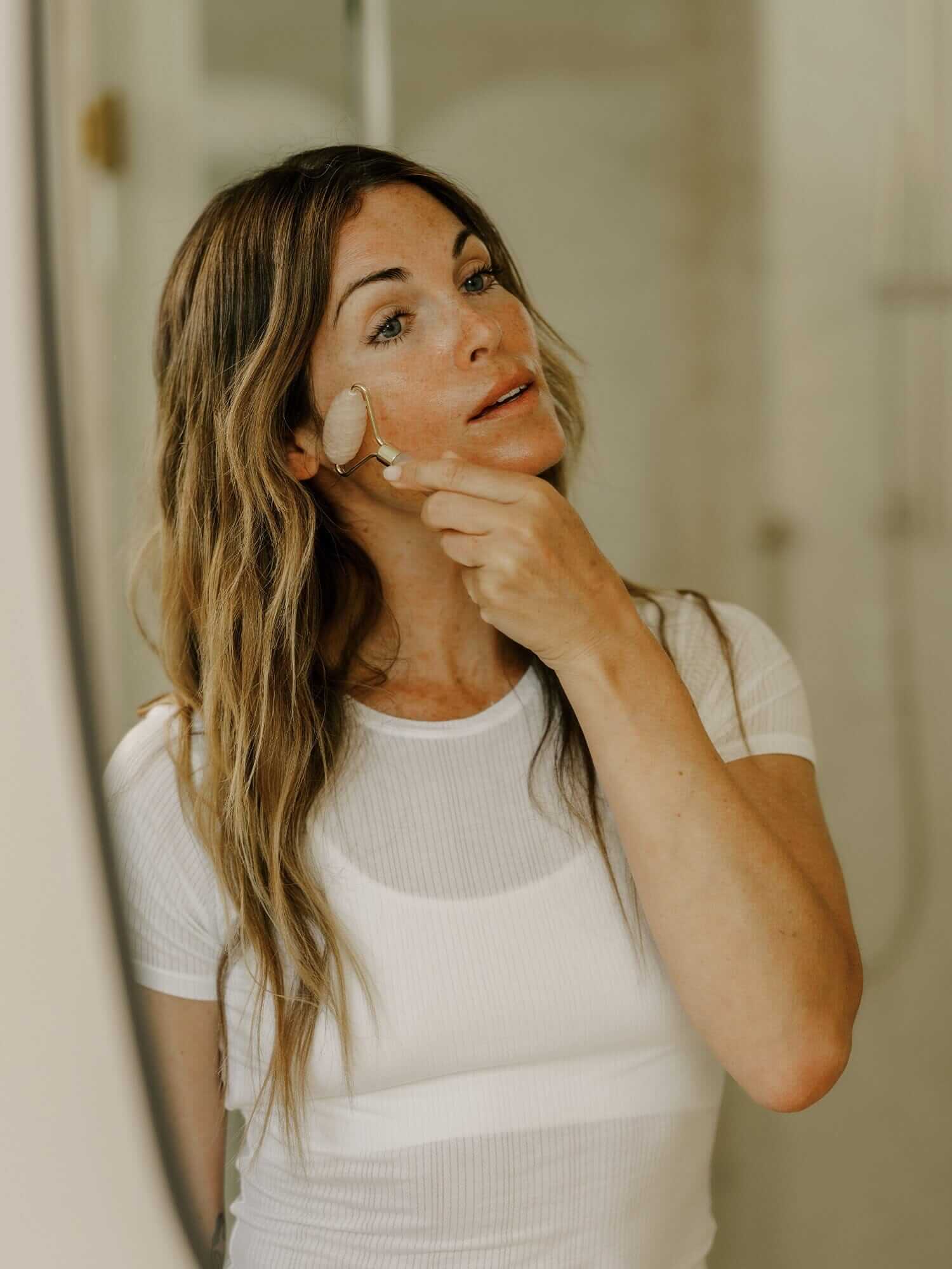 A woman in a white shirt uses a facial roller on her cheek while looking into a mirror in a bathroom.