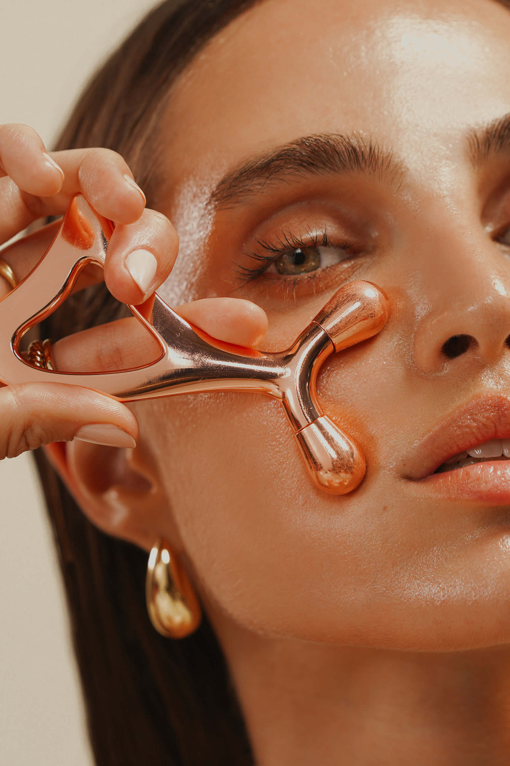 A woman uses a metallic facial massage tool on her cheek; her skin appears dewy and she wears gold hoop earrings.
