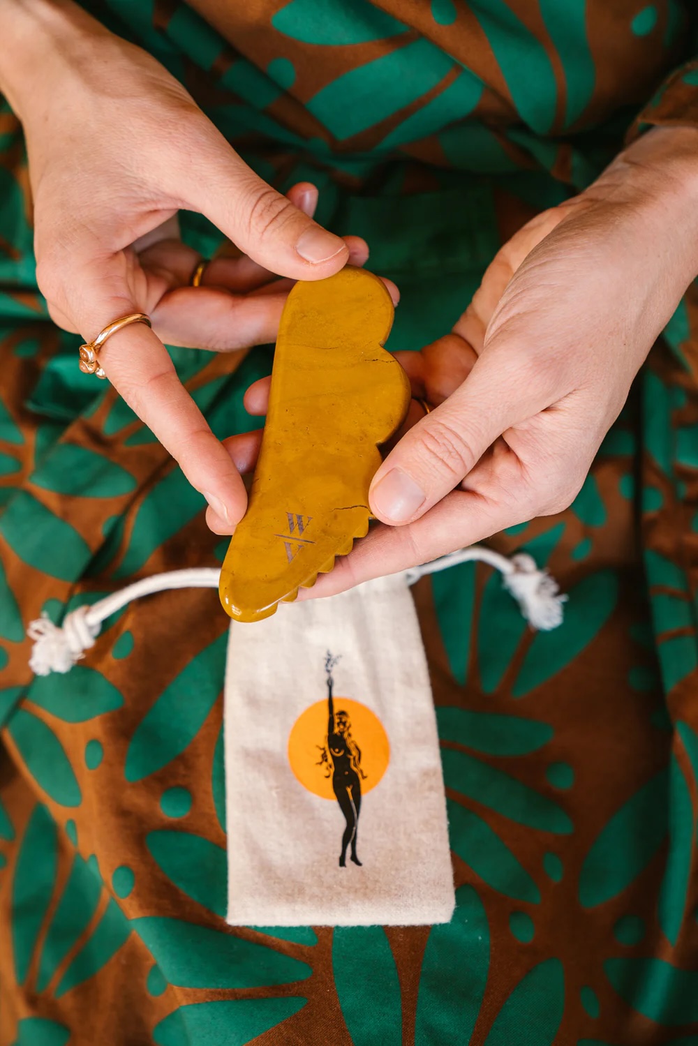 A person holds a yellow jade gua sha tool above a drawstring pouch with an illustration of a woman, against a patterned dress background.