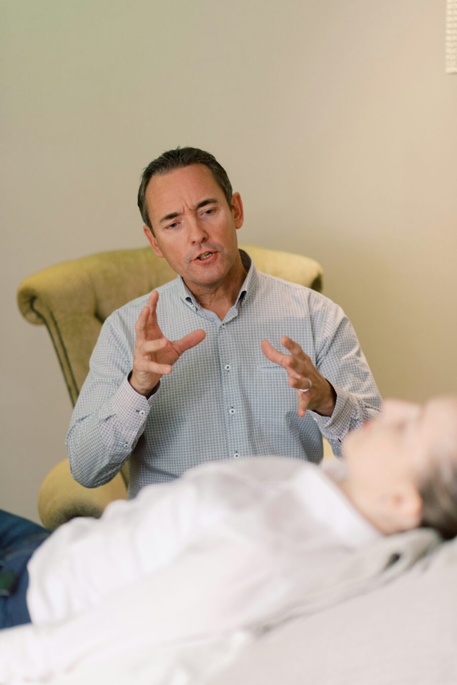 A man in a collared shirt speaks and gestures with his hands while seated beside a reclining person in a light-colored room.