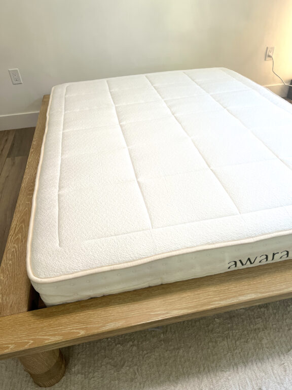 A neatly made bed with a white mattress labeled "awara" on a light wooden bed frame, positioned in a minimally furnished room with light walls and wood flooring.
