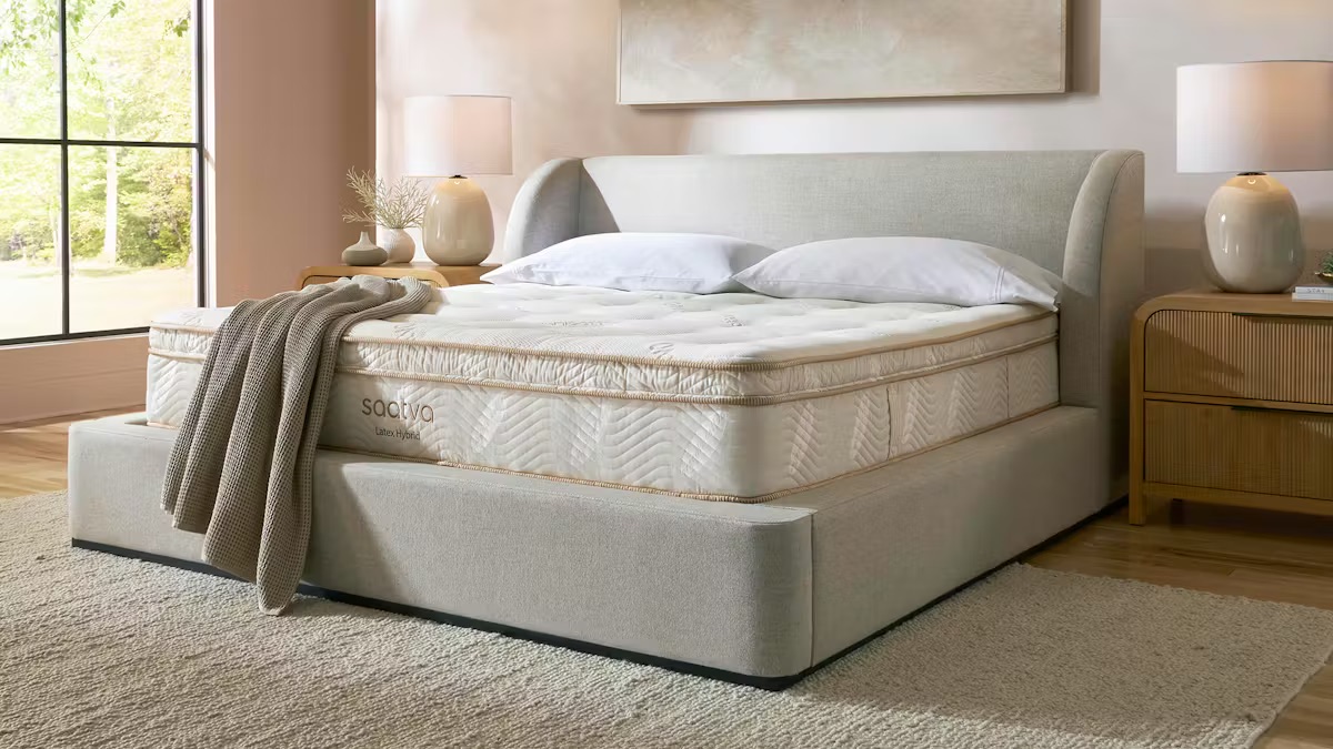 A neatly made bed with a light gray upholstered frame and headboard, topped with a thick mattress, two pillows, and a beige throw blanket in a softly lit bedroom.