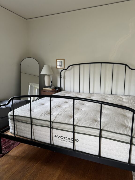 A bedroom with a black metal bed frame, an Avocado Green Mattress, a nightstand with a lamp, and a tall mirror in the corner.