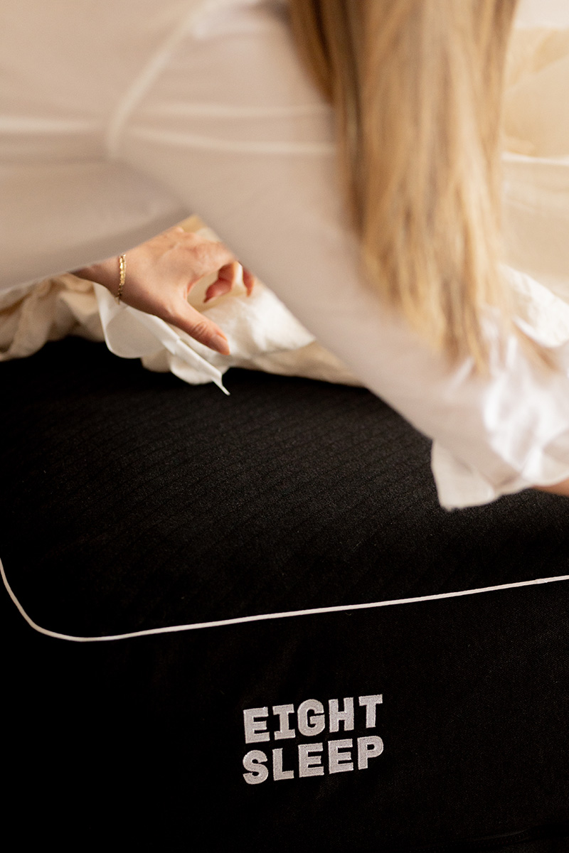 A person in a white shirt leans over a black mattress with "EIGHT SLEEP" written on its side in white letters.