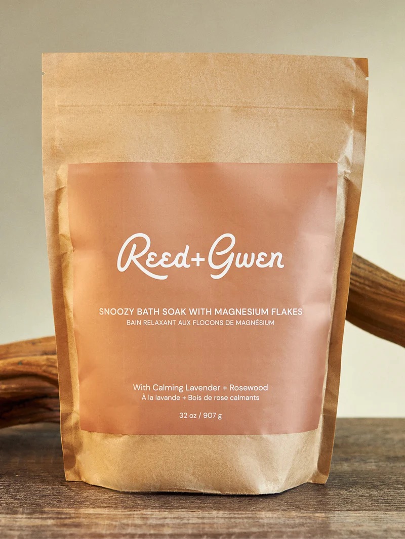 A brown bag of Reed + Gwen Snoozy Bath Soak with Magnesium Flakes, featuring lavender and rosewood, 32 oz size, displayed upright on a wooden surface.