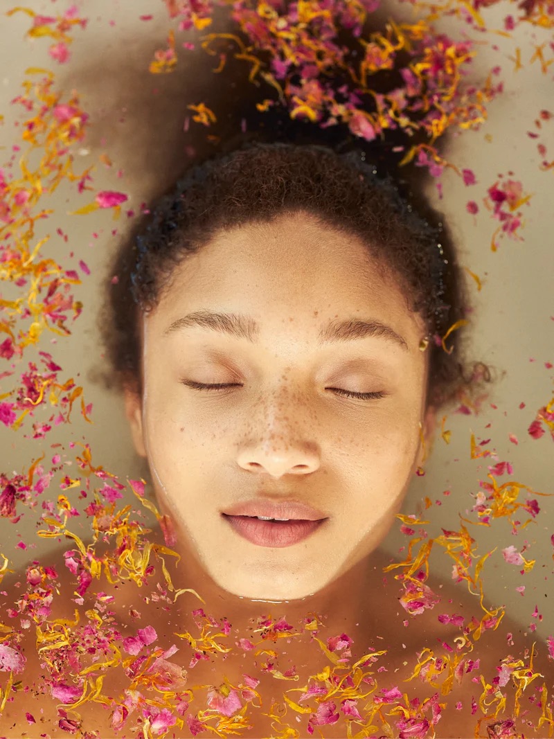 A person with closed eyes is partially submerged in water, surrounded by floating colorful flower petals.