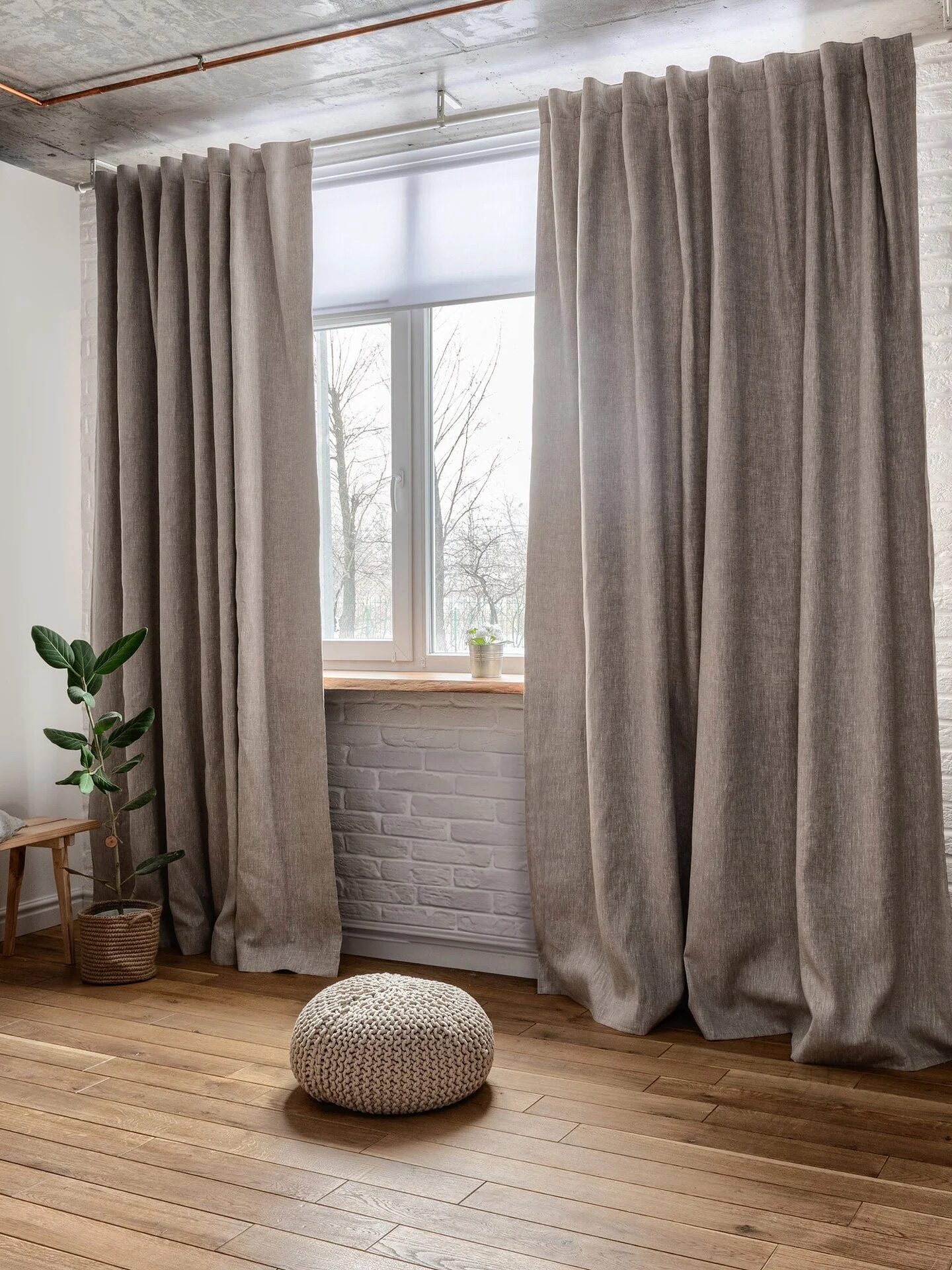 A room with wooden floors, beige curtains framing a window, a potted plant on a bench, a round cushion on the floor, and exposed copper pipes on a white brick wall.