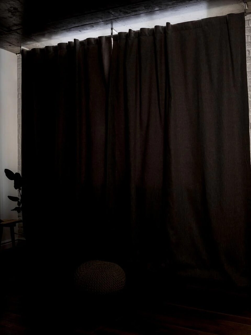 A dark room with closed black curtains covering a window, a stool, a round cushion, and a potted plant are partially visible in the dim light.