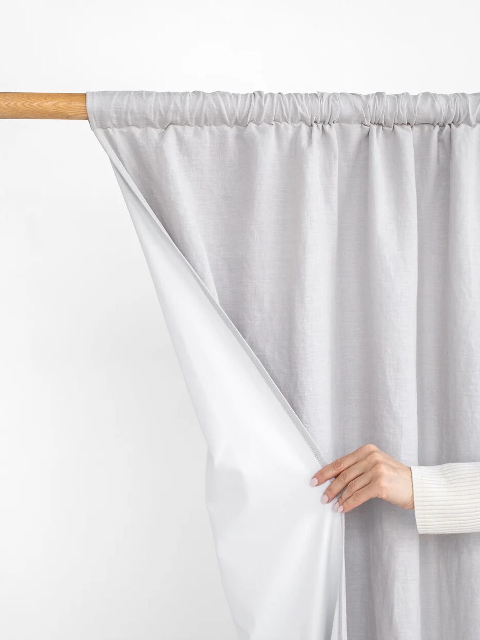 A hand pulls back a gray curtain to reveal a white inner lining, both hanging on a wooden rod against a plain white background.