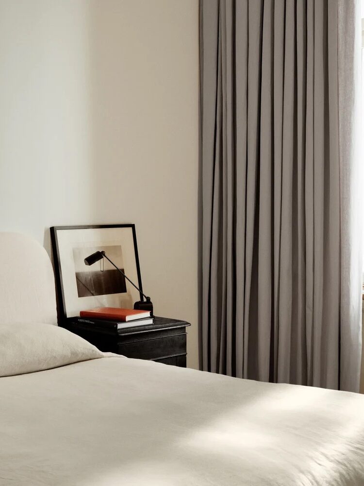 A minimalist bedroom with a neatly made bed, a small black nightstand holding books, a framed picture, a lamp, and long gray curtains by a sunlit window.