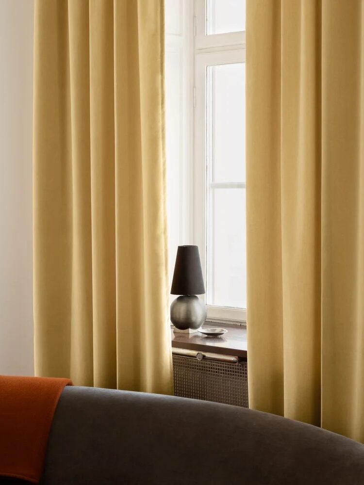 A window with closed yellow curtains, a small black lamp, and a round silver base on the windowsill; a gray sofa with an orange throw is in the foreground.