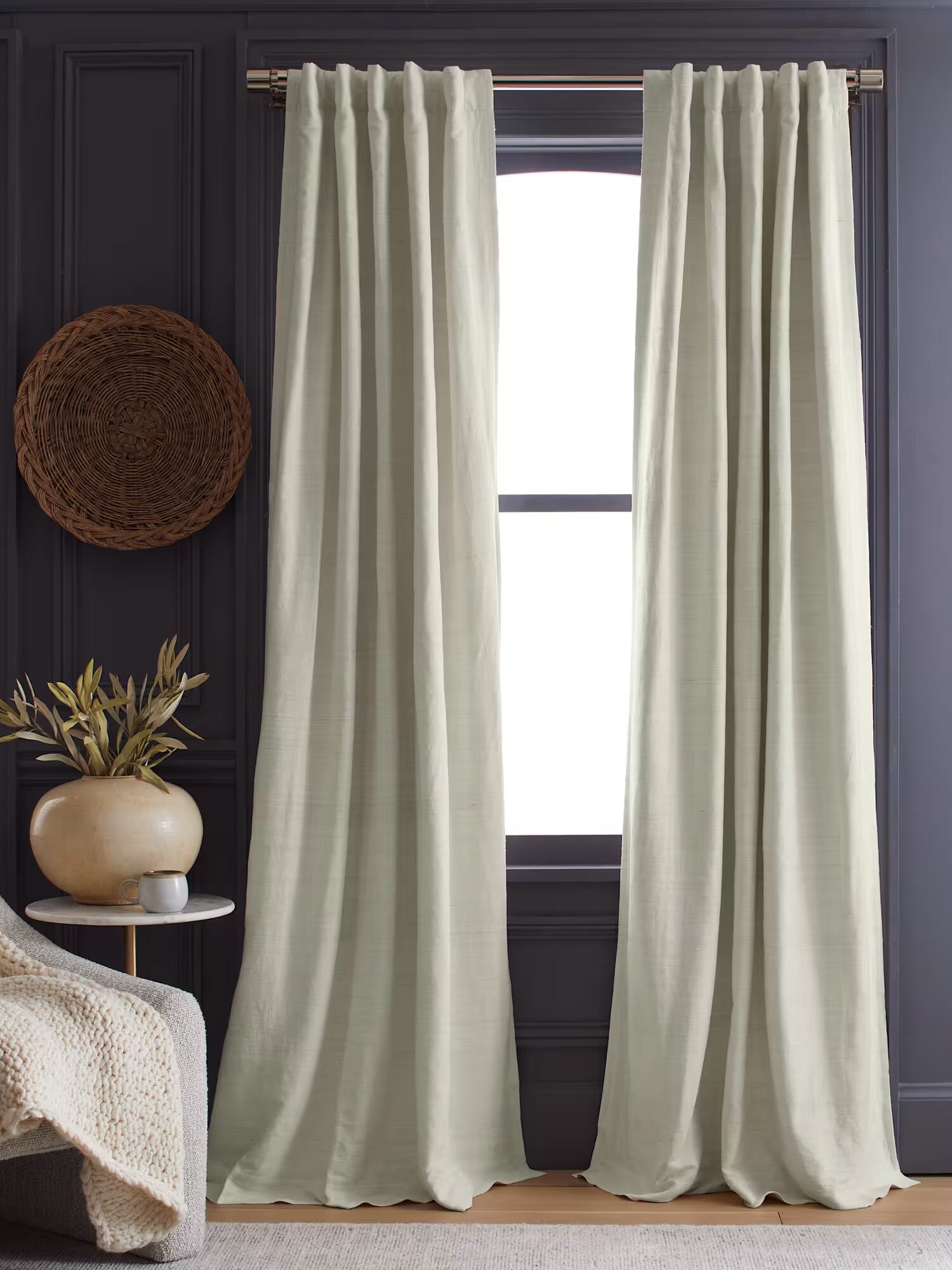 A pair of light beige curtains hang on a window with a brass curtain rod in a dark room featuring a woven wall decoration and a potted plant on a side table.