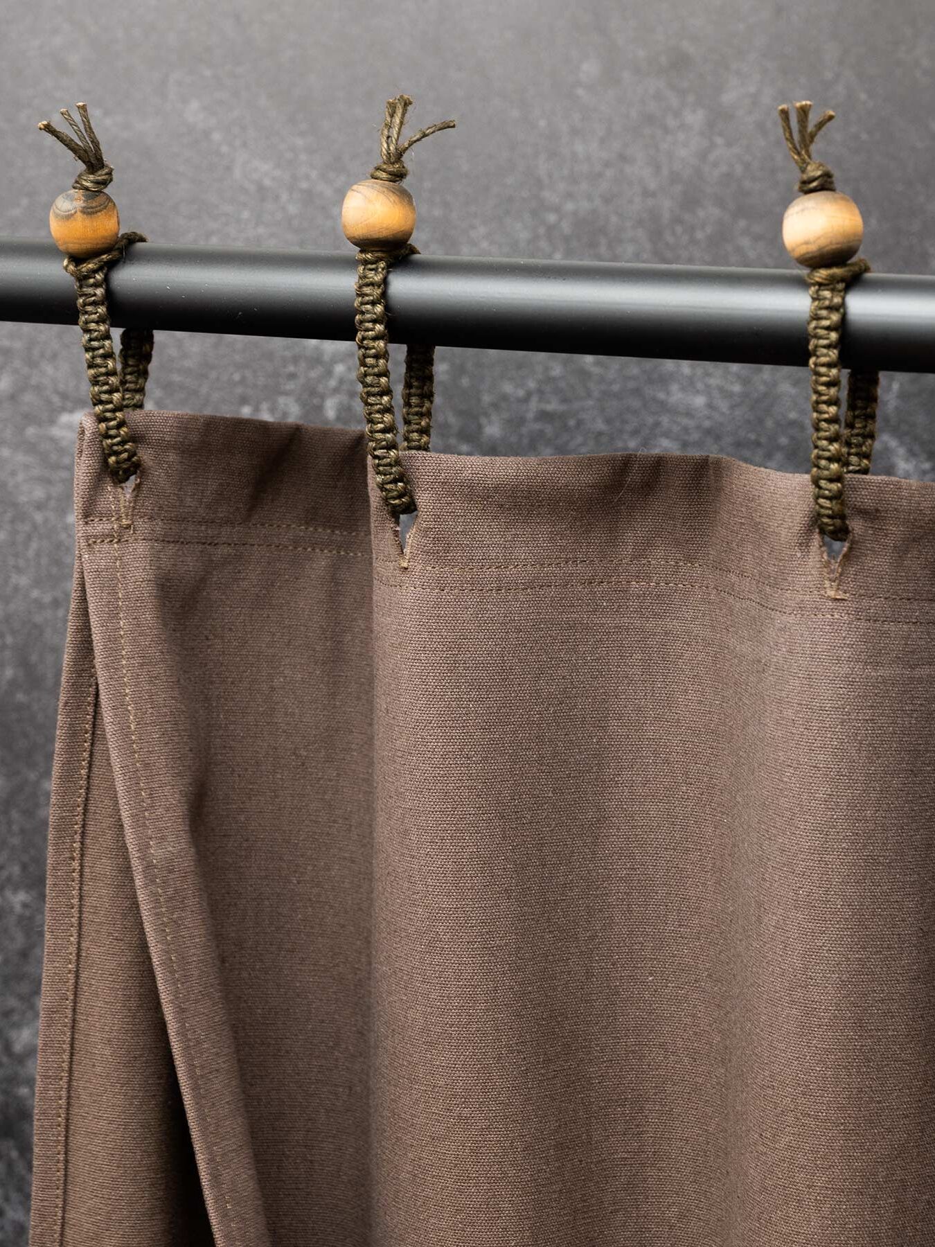 A brown curtain is hung from a black rod using braided loops with wooden beads against a textured dark background.