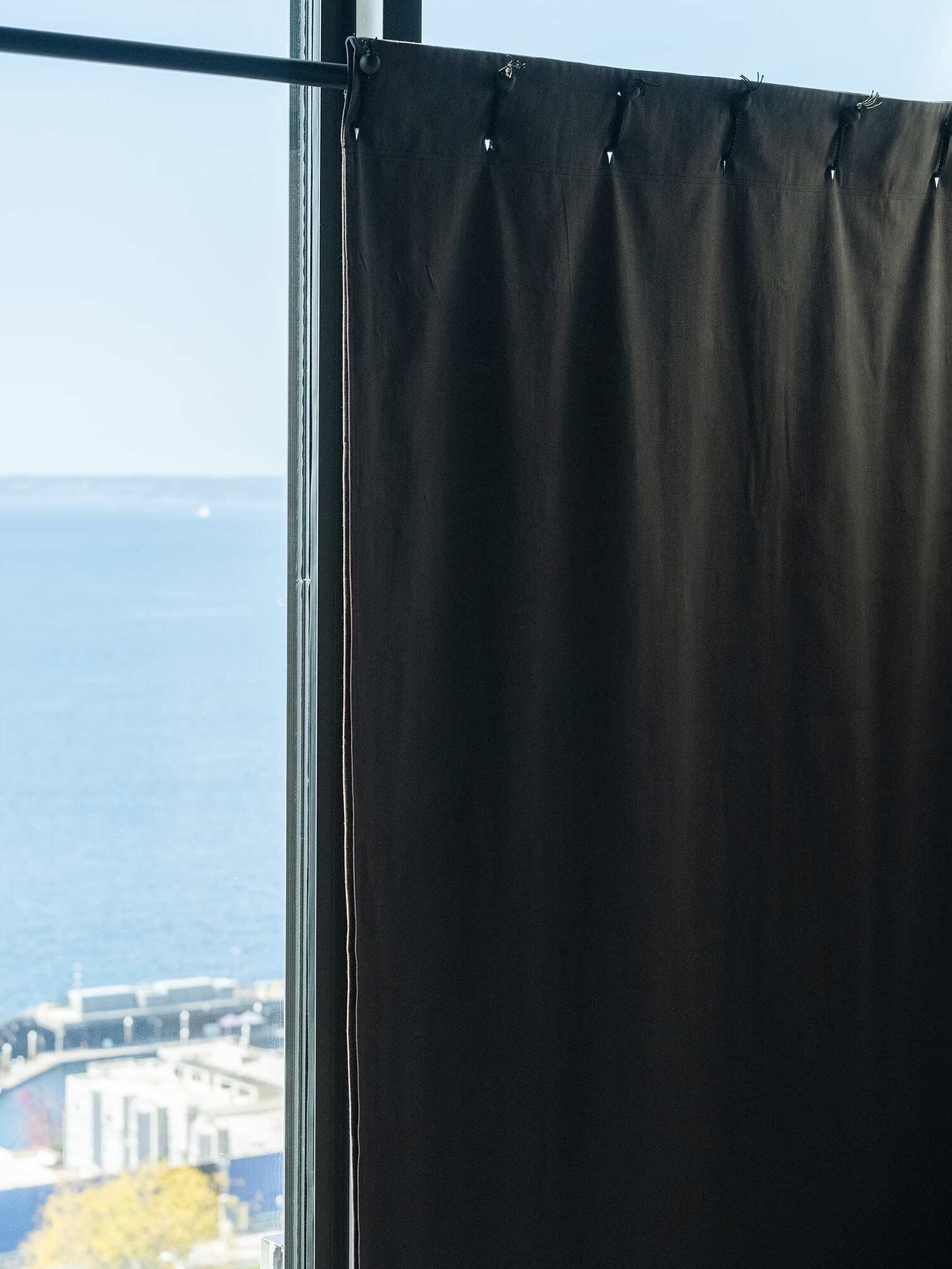 A dark curtain partially covers a large window with a view of the ocean and a section of buildings below.