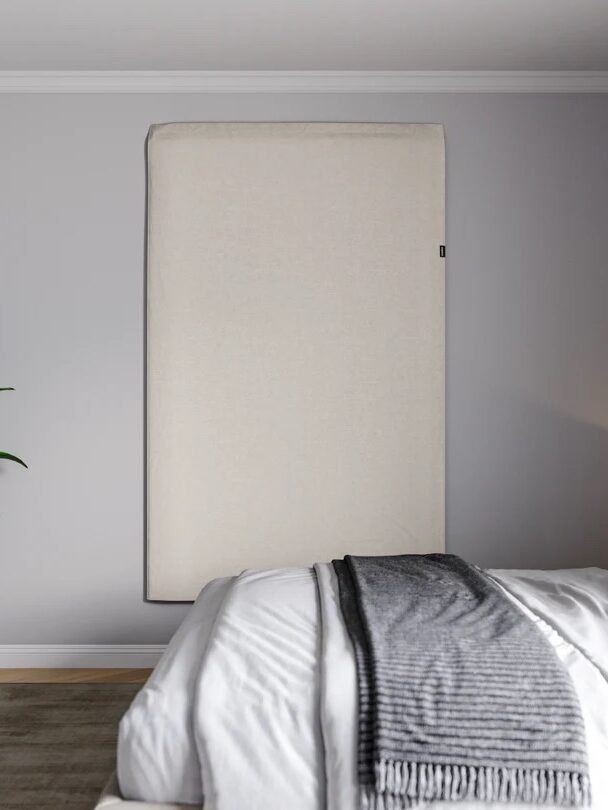 A large rectangular beige panel is mounted vertically on a light gray bedroom wall above a bed with white bedding and a gray throw blanket. A green plant is in the corner.
